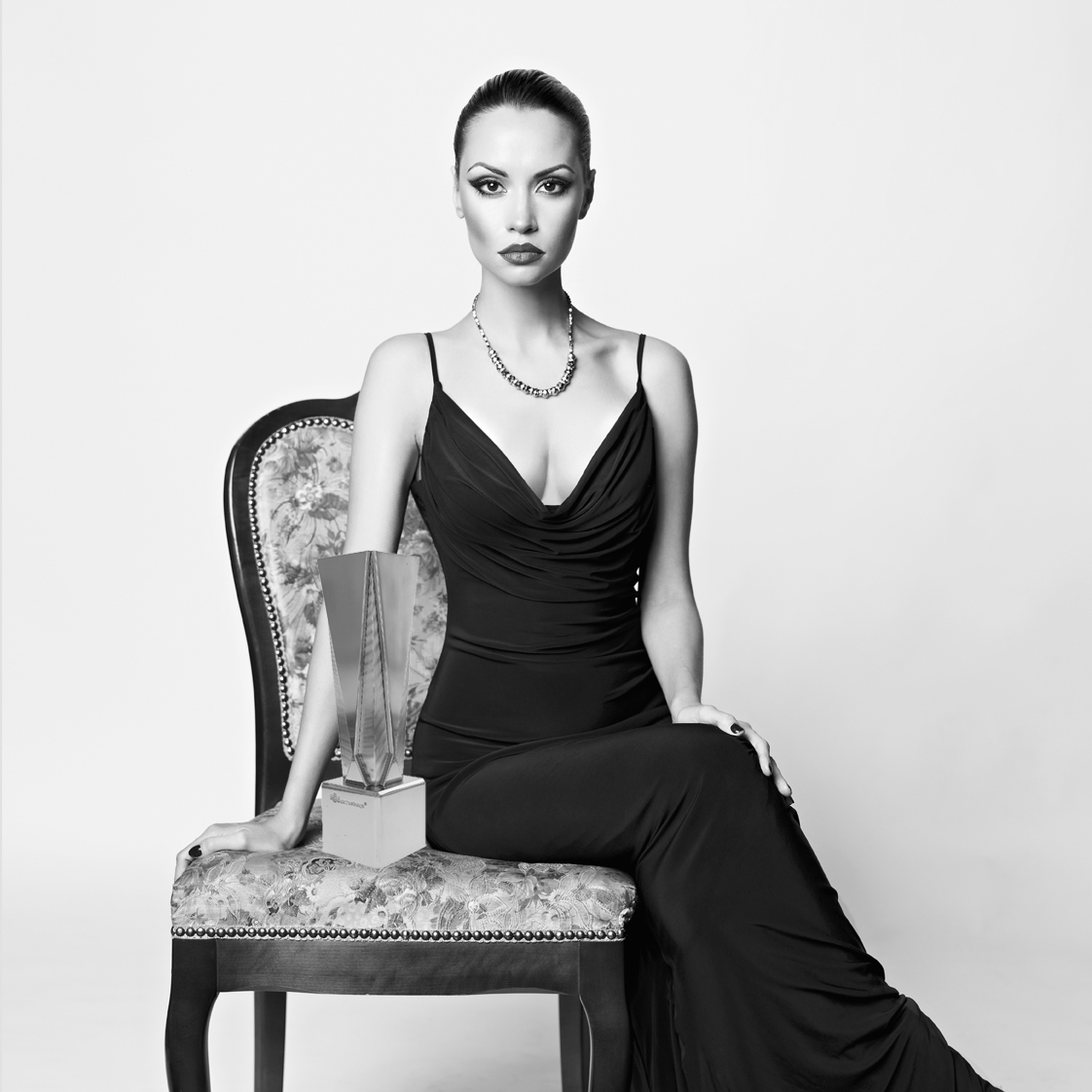 Model in black evening gown, sitting on a damask side chair with the Gatsby Silver Small award from the Exclusives Line of trophies.