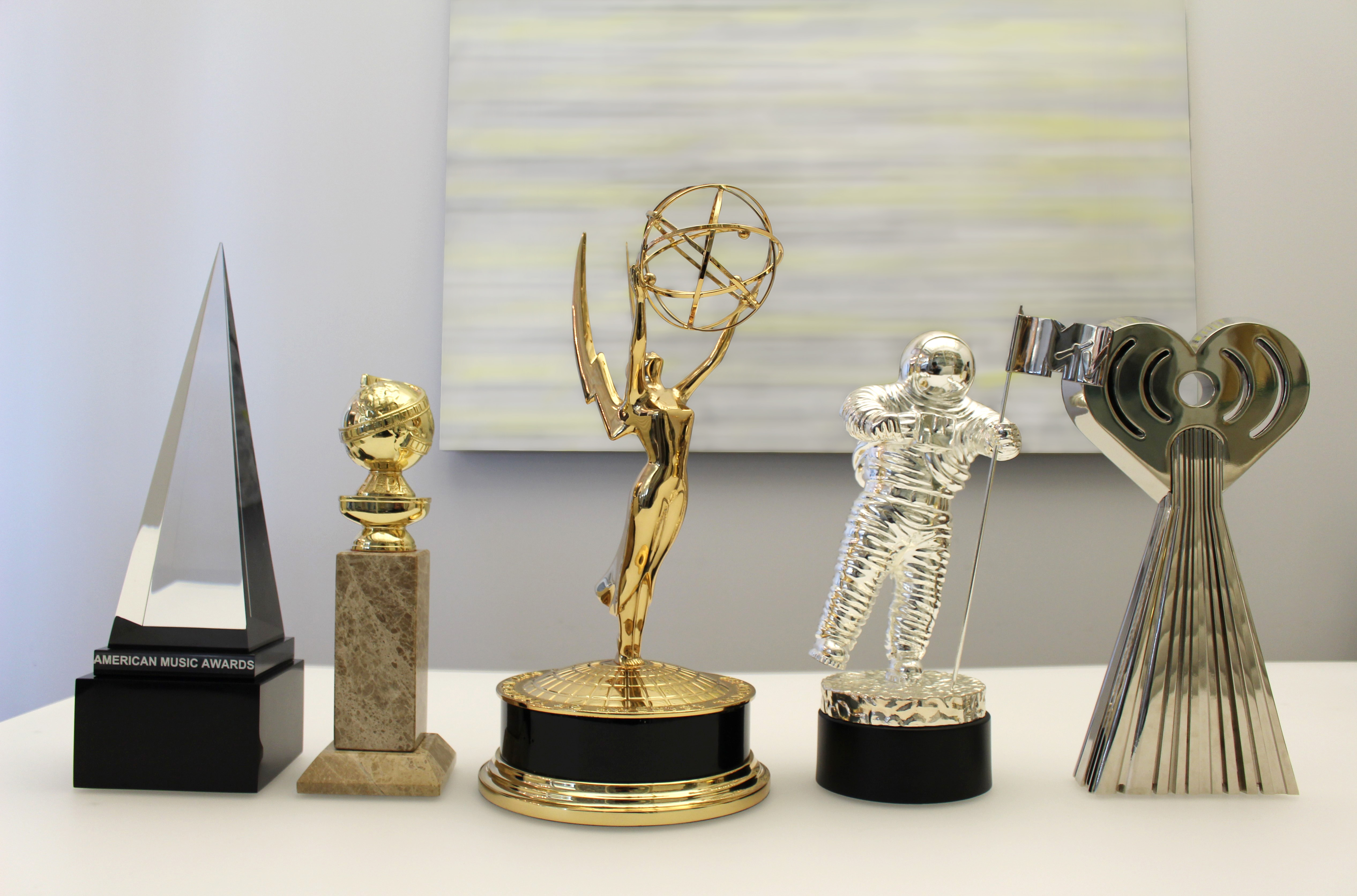 5 famous awards by Society Awards lined up on white table. From left to right: AMA, Golden Globe, Emmy, MTV VMA, iHeartRadio Music Award.