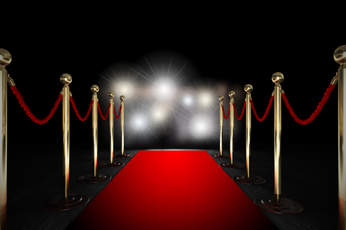 Red carpet with velvet ropes on either side and flashing lights of cameras in the distance.